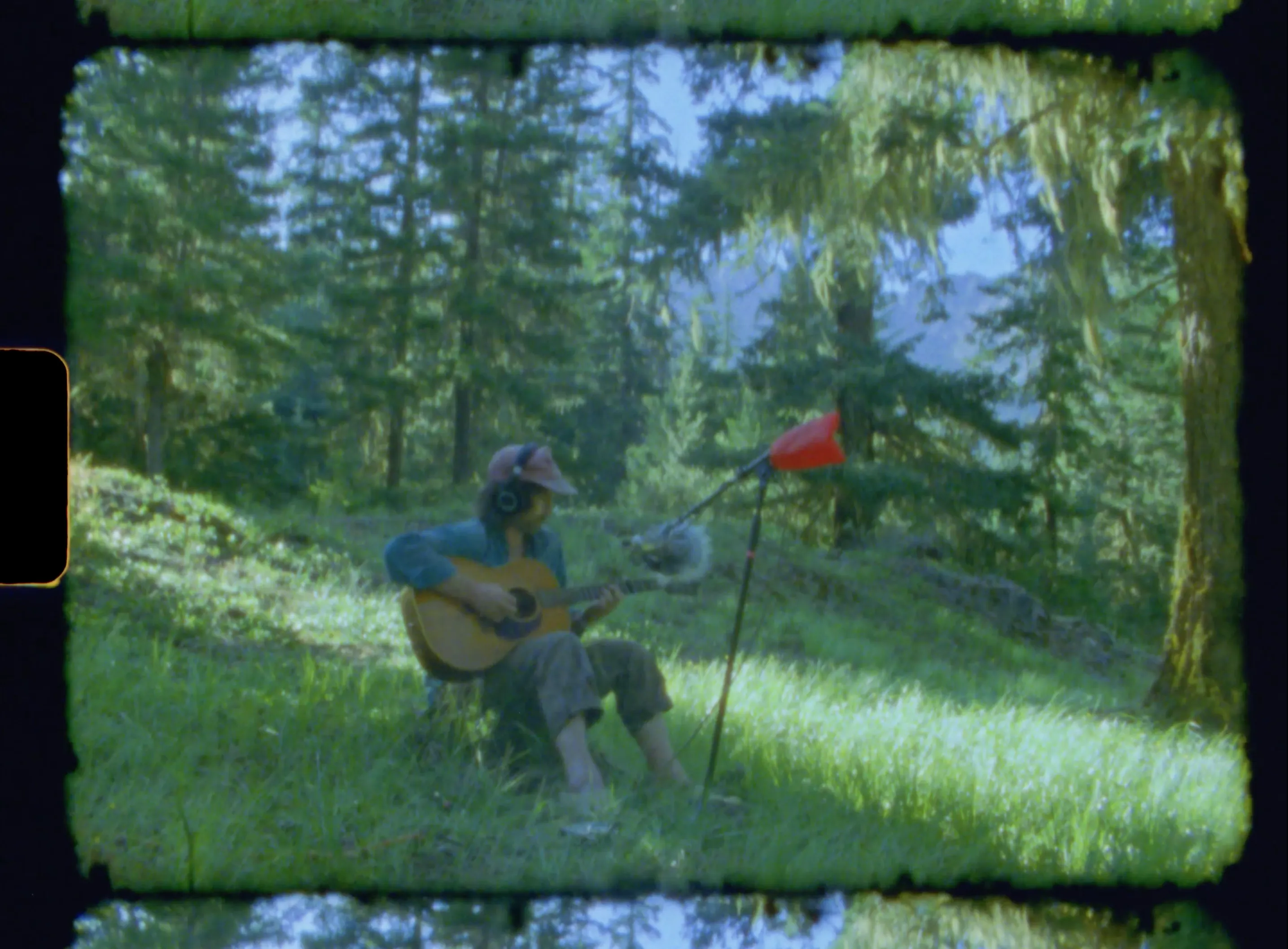 A guitarist in headphones and a pink cap plays acoustic guitar on a mossy forest slope, a windscreened microphone on a stand in front of him